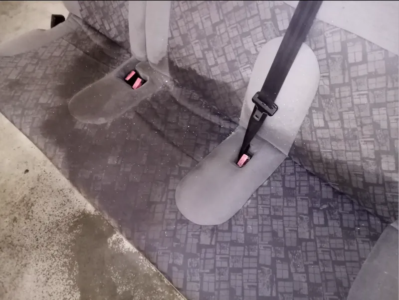 Zadní sedací lavice vymontovaná z vozu a postavená na podlaze dílny, na šedém vzorovaném potahu jsou tmavé mokré skvrny a zašlá místa kolem spon bezpečnostních pásů.