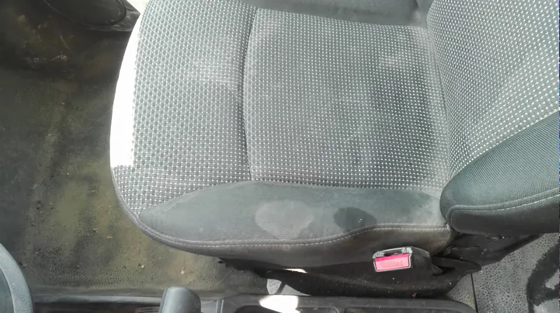 Sedák předního sedadla zblízka, na tmavém vzorovaném potahu jsou rozlité skvrny a zašlá místa, vlevo je vidět podlaha u dveří.