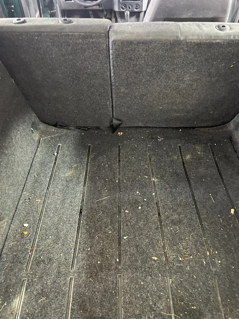 Zavazadlový prostor kombi před vyčištěním: na tmavém koberci a kolejnicích drobky, stébla a další nečistoty.